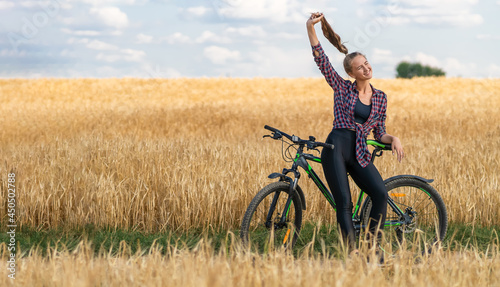 Beautiful young woman has her hair pulled up in a ponytail and is smiling while sitting on a bicycle in the middle of a field with triticale on summer day.Concept of a rural lifestyle, sports.Banner