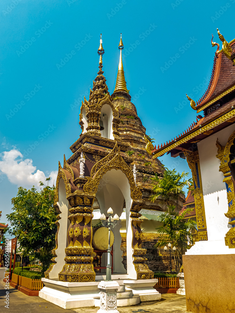 Traditional Norther Thai style bell tower, decorated with small golden umbrella on the top, in front of an old pagoda in the Buddhist temple under blue sky