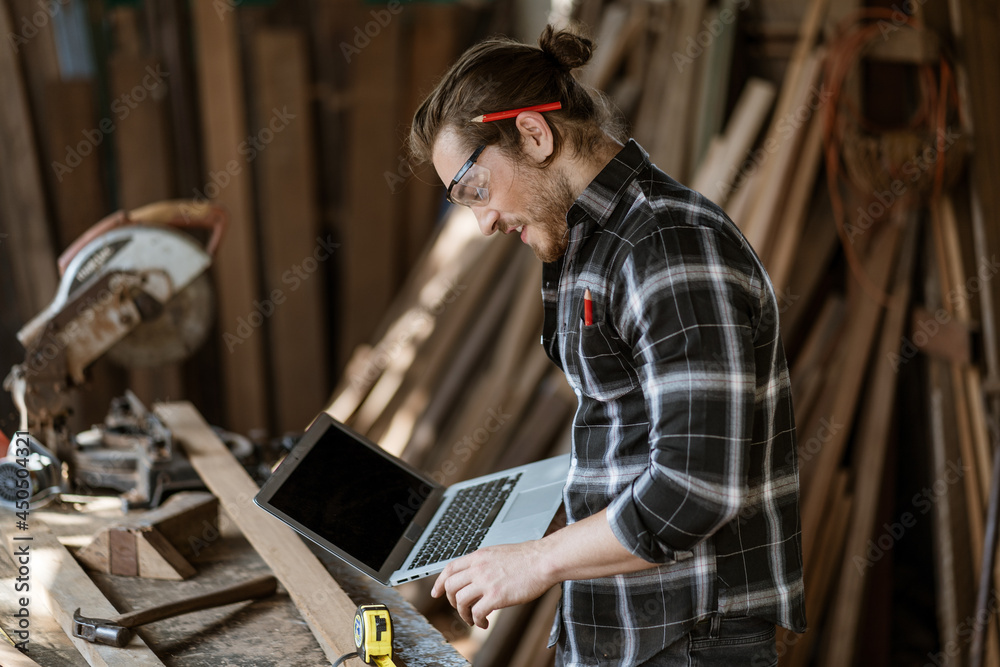 young hipster carpenter man holding computer laptop looking and checking wood in workshop . craftsman wearing safety glasses choosing plank young hipster carpenter man holding computer laptop looking and checking wood in workshop . craftsman wearing safety glasses choosing plank