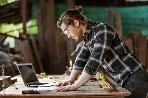 young hipster carpenter man working with computer laptop in workshop . craftsman wearing safety glasses checking order of clients or learning online
