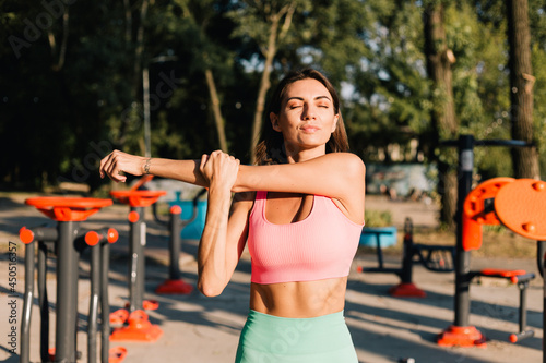 Sporty woman in fitting sport wear at sunset at outdoor sports ground stretching her arms shoulders before after workout