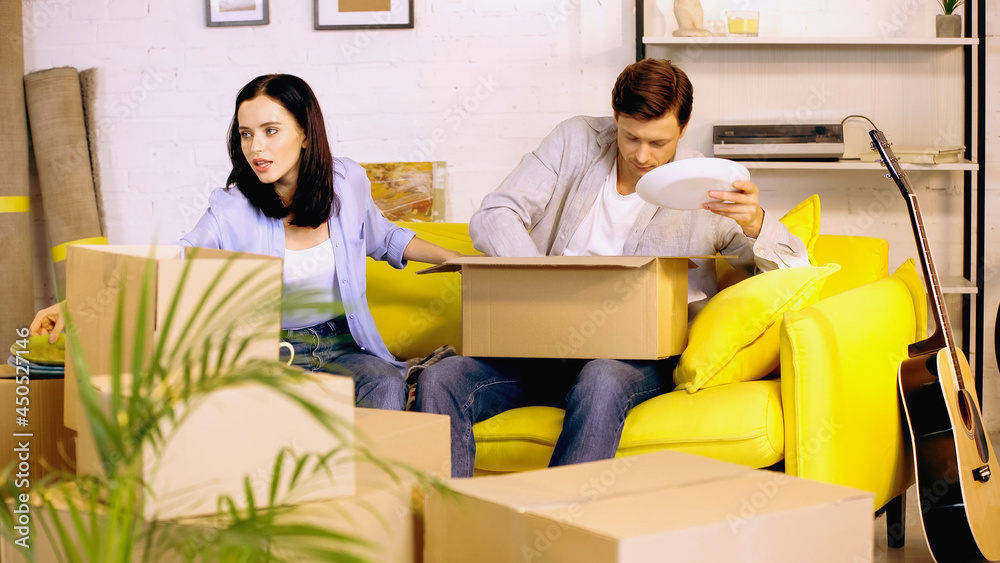 Couple packing plates in carton boxes on couch Couple packing plates in carton boxes on couch