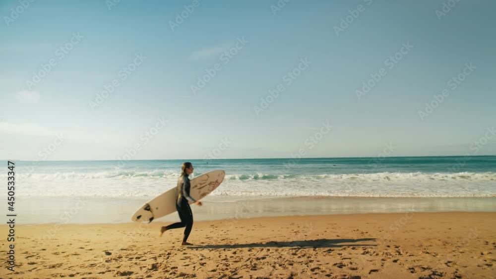 Cinematic warm beach shot of young fit athletic woman in wetsuit run on edge of water and sandy beach towards ocean. Female surfer carry surfboard to waves. California surfing vibes lifestyle Cinematic warm beach shot of young fit athletic woman in wetsuit run on edge of water and sandy beach towards ocean. Female surfer carry surfboard to waves. California surfing vibes lifestyle