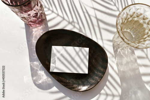 Summer mockup tableware scene.  Empty blank on ceramic plate, cocktail glass on sunlit background. Palm shadow.  Mock invitation card for wedding, business or menu. Eco minimal wabi sabi style