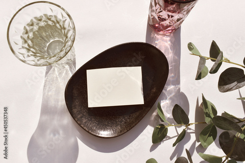 Eco minimal still life scene.  Empty blank on ceramic plate, cocktail glass and eucalyptus leaves on neutral background. Mockup invitation card for wedding, business or menu. Sunlight, long shadow