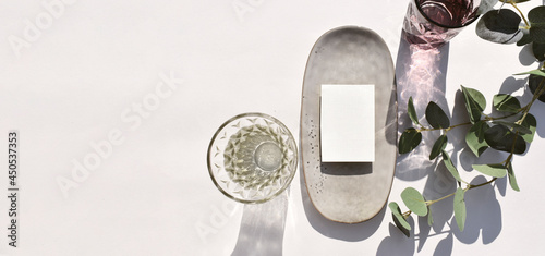 Mock up empty blank invitation card, ceramic plate cocktail glass and eucalyptus leaves on sunlit background. Summer mockup table setting  for wedding, business or menu. Long shadows