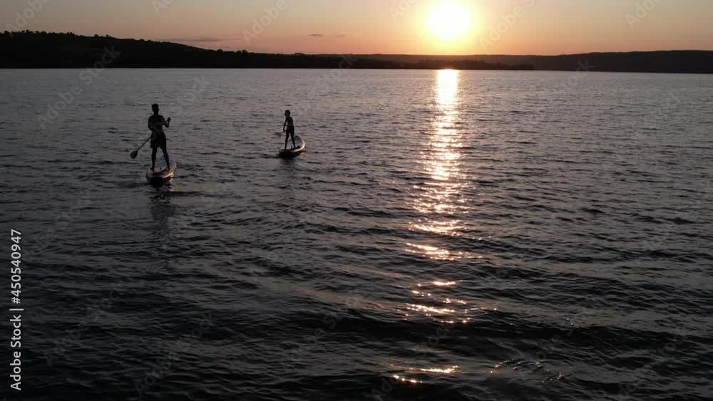 Silhouette of people standing on SUP board and paddling at sunset, waves on water. Aerial view. Beautiful landscape. Healthy lifestyle. Golden hour. Fitness concept Silhouette of people standing on SUP board and paddling at sunset, waves on water. Aerial view. Beautiful landscape. Healthy lifestyle. Golden hour. Fitness concept