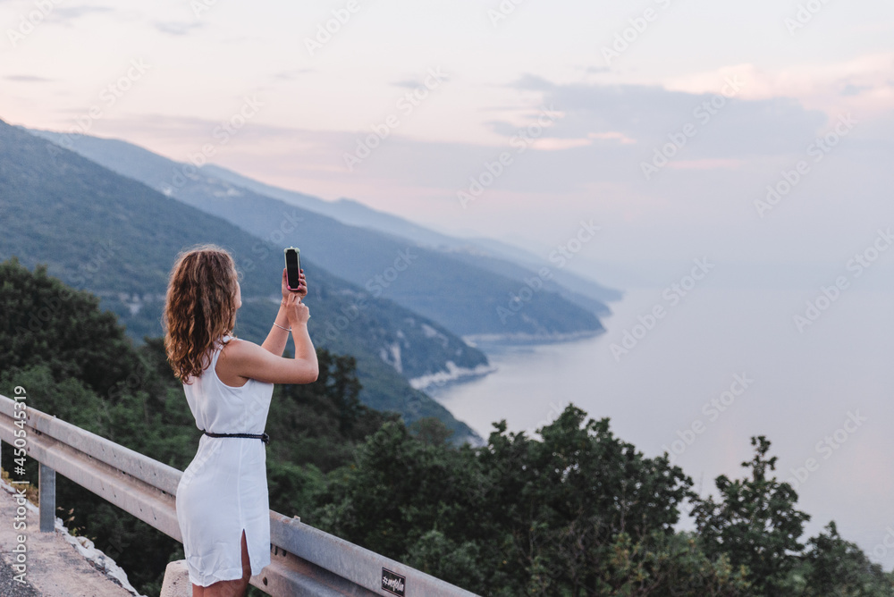 Naklejka premium stop on the road woman taking picture on the phone of sunset above the sea