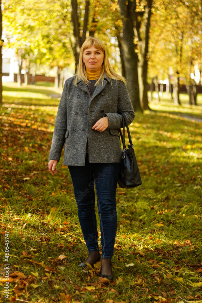 Defocus serious blond 40s woman standing in yellow autumn park. Happy beautiful lady. Women wearing grey coat, pullover, yellow turtleneck. Fall park, leaves. Overall vertical portrait. Out of focus