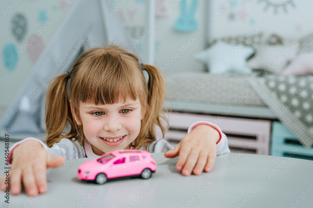 Beautiful little girl wearing pajamas playing with pink toy car in the kids room