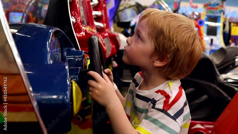 Little happy blonde caucasian boy playing driving game on modern slot machine in amusement park. Children activity. Urban family lifestyle. True feelings. Emotional male child riding a lot.