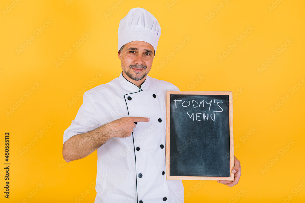 Chef cook wearing white kitchen hat and jacket holding a blackboard that says 'Today's menu', on yellow background Chef cook wearing white kitchen hat and jacket holding a blackboard that says 'Today's menu', on yellow background