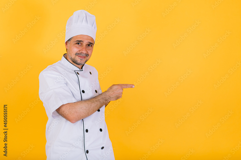 Chef cook wearing white kitchen cap and jacket pointing to the right with his hands, on yellow background Chef cook wearing white kitchen cap and jacket pointing to the right with his hands, on yellow background