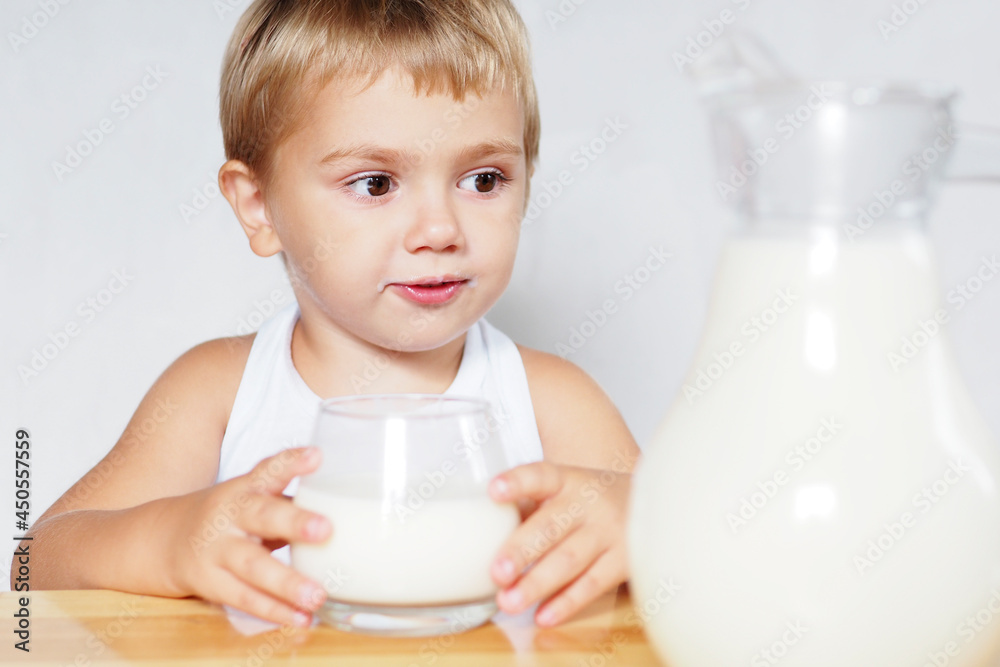smiling blond boy with brown eyes drinks milk from a glass at a wooden table on a white background... smiling blond boy with brown eyes drinks milk from a glass at a wooden table on a white background...