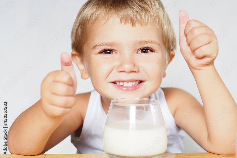a smiling blond boy with brown eyes drinks milk from a glass at a wooden table and shows his class with his hands... a smiling blond boy with brown eyes drinks milk from a glass at a wooden table and shows his class with his hands...