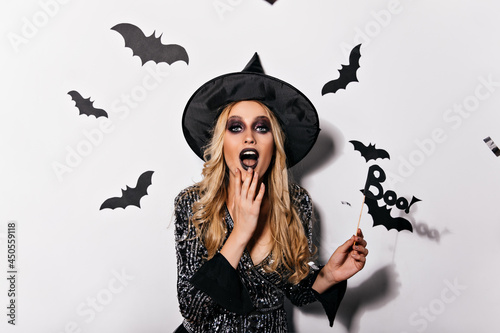 Interested blonde woman in witch costume playfully posing on white background. Studio photo of female vampire surrounded by bats.