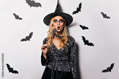Shocked european girl in halloween costume looking to camera. Adorable young witch in black attire posing with bats.