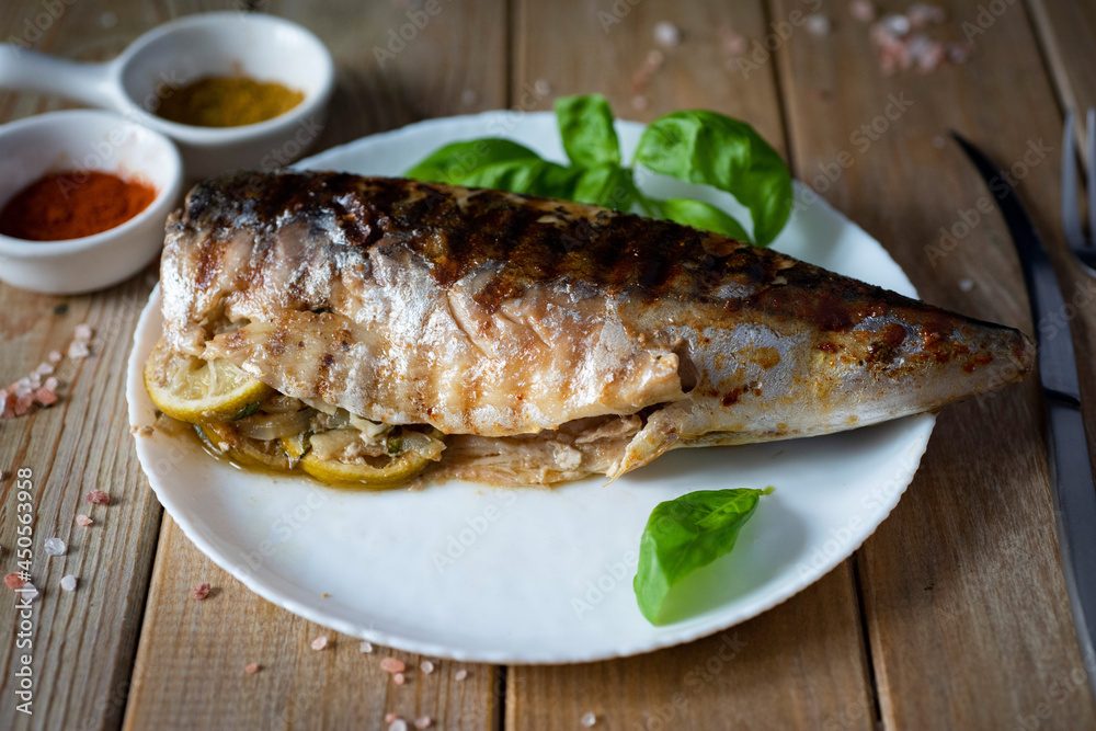 Fish dish: grilled mackerel stuffed with vegetables and lemon on a white plate on a wooden table. Close-up Fish dish: grilled mackerel stuffed with vegetables and lemon on a white plate on a wooden table. Close-up