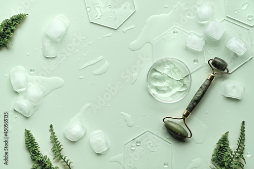 Moisturizer, green jade face roller with exotic fern leaves. Mint green background with moisturizer in chemical glass petri dish and ice. Minimal geometric flat lay with hexagones. Monochrome look.