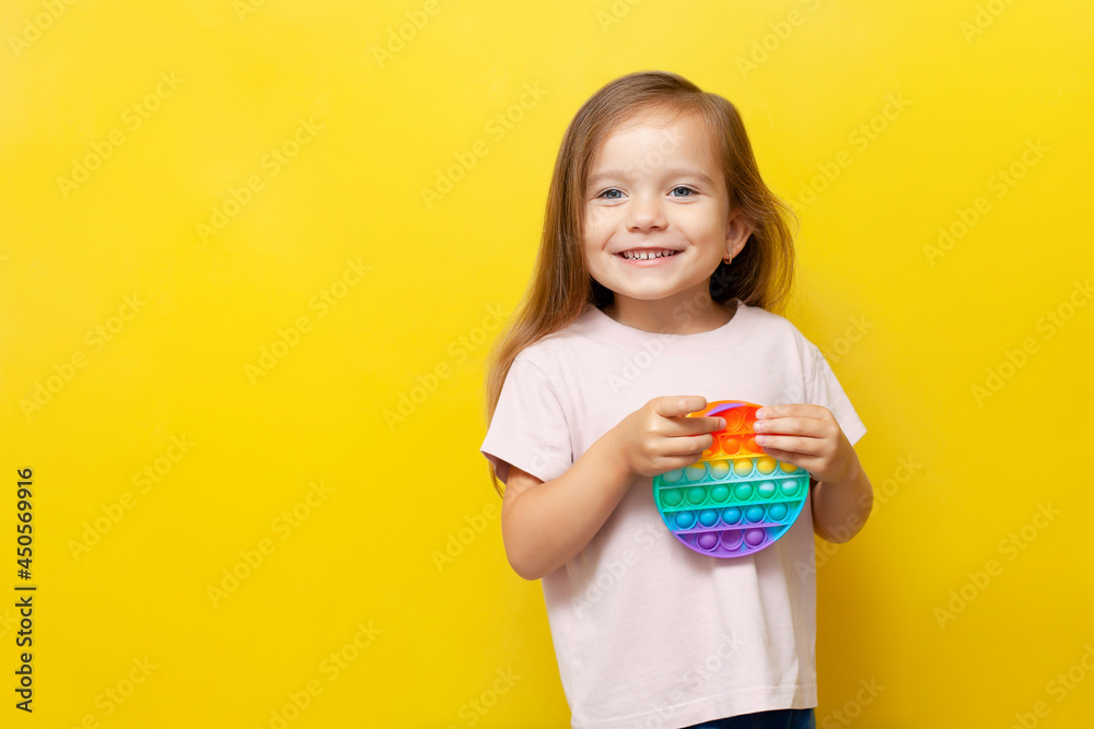 A beautiful cute girl holds a colored pop it toy in her hands and smiles. Yellow background. Anti-stress, emotions, good mood. A place for text. Trend