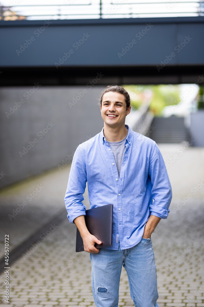 Portrait of young man holding laptop computer while standing in a city area Portrait of young man holding laptop computer while standing in a city area