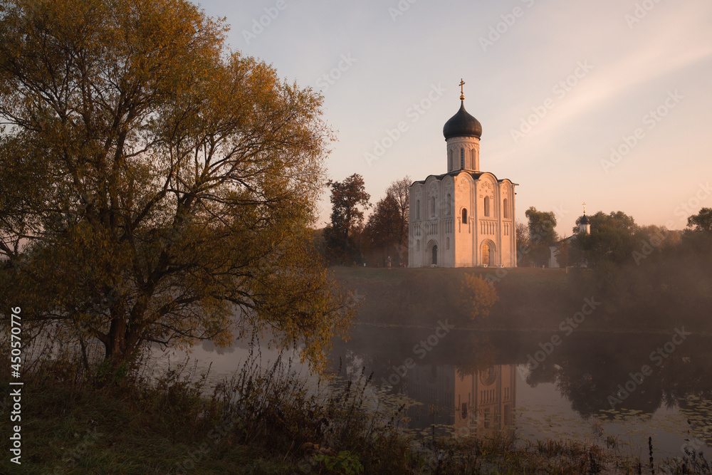 Old Russian Orthodox church lit by the rising sun above autumn forest in light mist reflecting in a pond with a big tree in the foreground