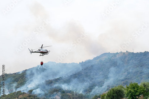 Fire helicopter dropping water from a bucket on a forest fire in the mountains. Forest fire is an environmental disaster. Deforestation.
