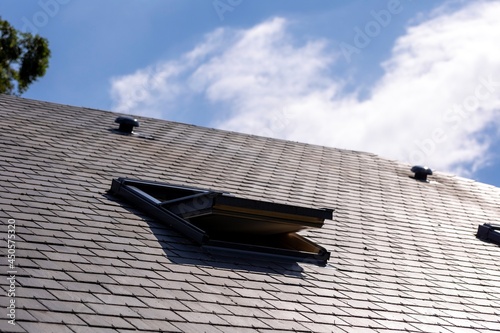 A portrait of an opened skylight window in a slate roof on a sunny day with a blue sky with white clouds. you can also see a ventilation vent on the rooftop.