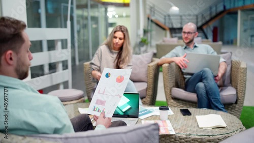 Startup team of three sitting in office lobby and discussing business strategy. Manager explaining market research stats and presenting printout to colleagues at meeting, focus on document photo