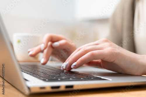 Close up photo of pretty woman hands typing on laptop keyboard. Female freelancer working from home. On the wooden desk notebook and cup of tea. Selective focus, blurred background. Side view.