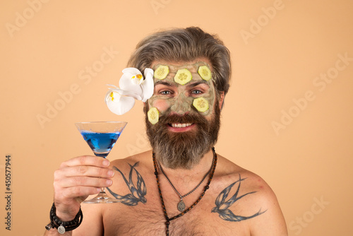 Funny man with clay mask and cucumber slices on face drink cocktail. Spa, dermatology, wellness and facial treatment concept. Man having cosmetic moisturizing mask, isolated studio background.