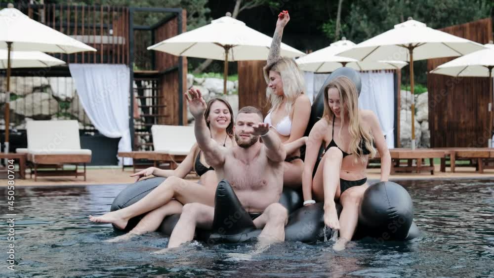 Cheerful girls and one guy playing with inflatable mattress floating in pool. Smiling friends enjoying summer vacation on inflatable swan flamingo. They are dance and smile