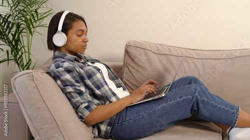 A young African-American woman in casual clothes works at home online with a laptop on her lap and listens to music in large white headphones. The concept of freelancing.
