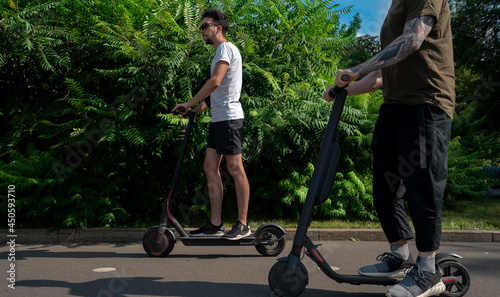 Two Friends Driving Electric Scooter In Park And Having Fun At Sunny Day.