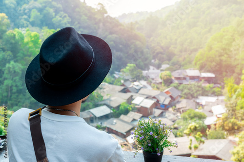 Fotografi Behind a young Asian man in a white shirt with a black hat sits on the balcony of a coffee shop overlooking a rural village below the mountains