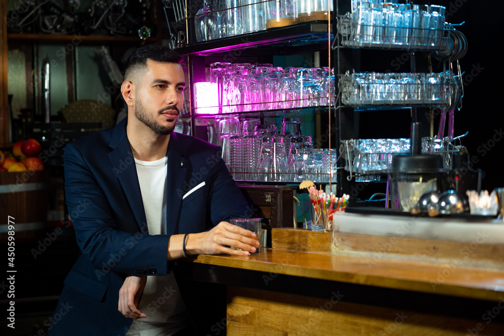 Handsome Caucasian man sitting at bar counter holding whiskey glass with ice cube and drinking alcoholic drink from barman in nightclub. Nightlife holiday party celebration and small business concept