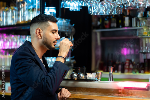 Handsome Caucasian man sitting at bar counter holding whiskey glass with ice cube and drinking alcoholic drink from barman in nightclub. Nightlife holiday party celebration and small business concept