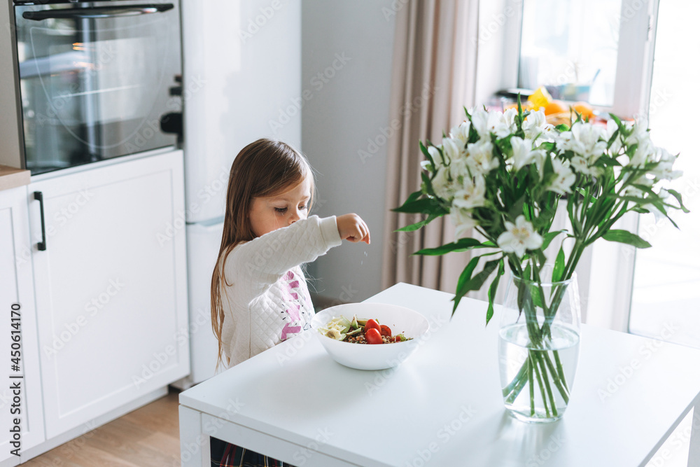Pretty little girl with long hair sprinkles salt on vegetable salad in kitchen with bright interior at home Pretty little girl with long hair sprinkles salt on vegetable salad in kitchen with bright interior at home