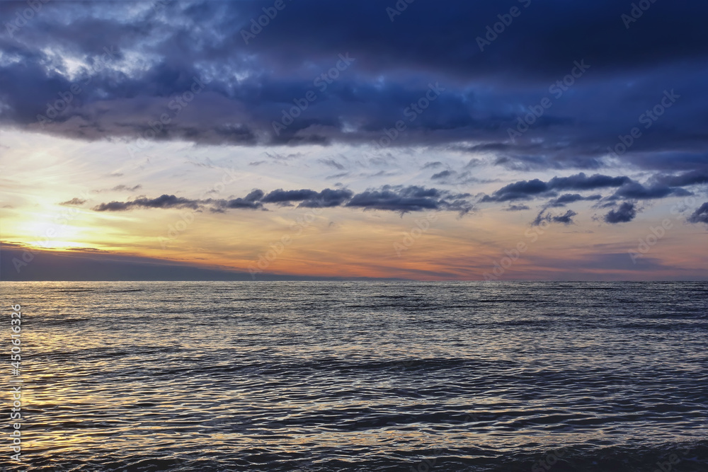 Sunset over the sea. There are dark blue clouds over the water. Small waves and ripples on the surface. The sky is highlighted in orange