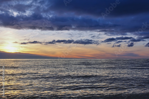 Sunset over the sea. There are dark blue clouds over the water. Small waves and ripples on the surface. The sky is highlighted in orange