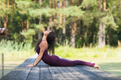 A woman in a burgundy overalls practicing yoga in the park, lying on a mat, performs the Bhujangasana exercise, the cobra pose photo