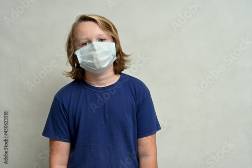 Young boy teenager in a medical mask with long hair in a blue t-shirt posing on a white background