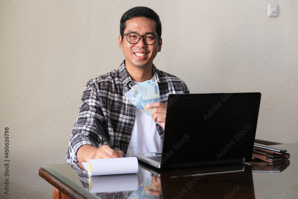 Adult Asian man smiling while holding paper money in front of laptop Adult Asian man smiling while holding paper money in front of laptop