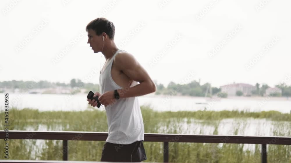 A side view of a muscular guy is running while listening the music on his mobile in the park outside A side view of a muscular guy is running while listening the music on his mobile in the park outside