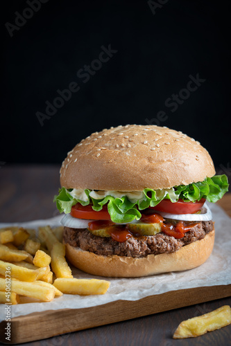 Juicy hamburger, also burger, made of cooked grilled patties of ground beef meat, lettuce,  tomato, onion, pickles inside fresh sliced bun served on wooden board with french fries on dark brown table