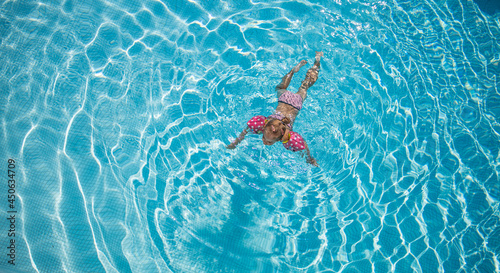Little girl playing in outdoor swimming pool water on summer vacation. Child learning to swim in outdoor pool luxury resort  top view. Cute toddler girl playing relaxing in turquoise water above view