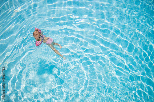 Little girl playing in outdoor swimming pool water on summer vacation. Child learning to swim in outdoor pool luxury resort, top view. Cute toddler girl playing relaxing in turquoise water above view