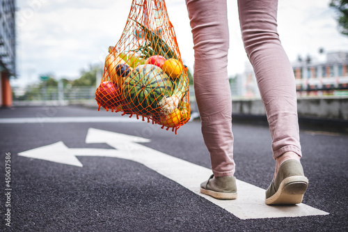 Choose the right direction for healthy and sustainable lifestyle with zero waste. Woman with reusable mesh bag full of fruit walking on the road from supermarket photo