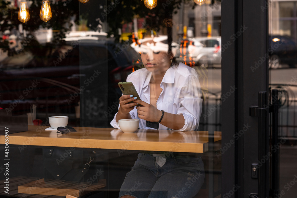 Woman at trendy cafe drinking coffee or tea and use smartphone. Young beautiful Caucasian model in white shirt and red lipstick. Reflections from windows. Shot through the window of fashion girl.