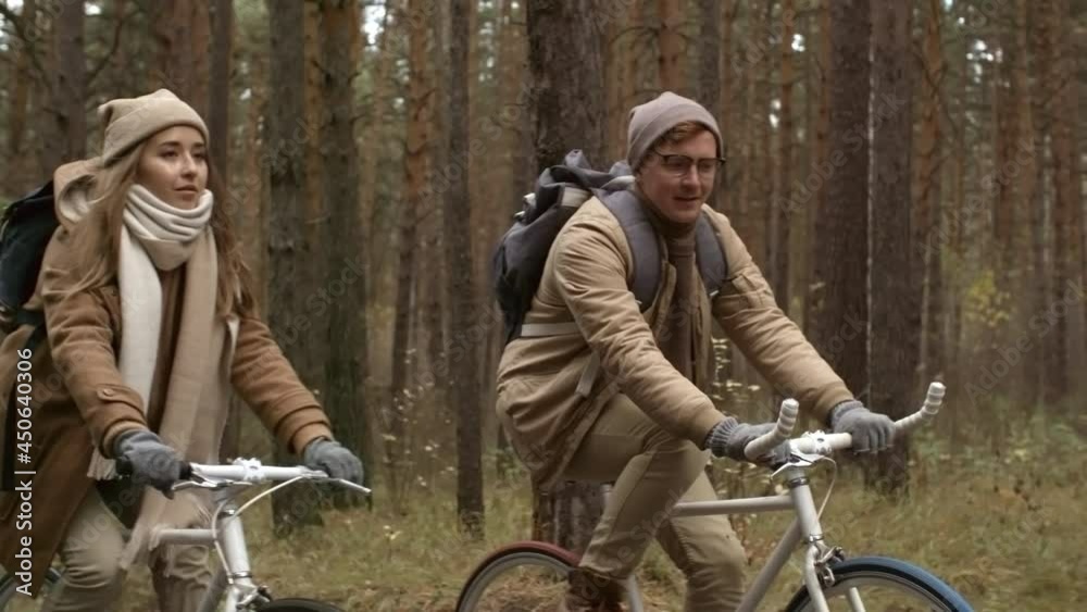 Tilt down shot of young man and woman in warm outerwear riding bikes and talking while cycling in autumn forest Tilt down shot of young man and woman in warm outerwear riding bikes and talking while cycling in autumn forest